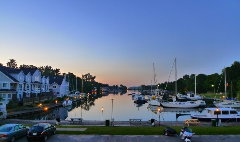 A waterfront at dusk, featuring sailboats and boats on the right side, and houses on the left side.