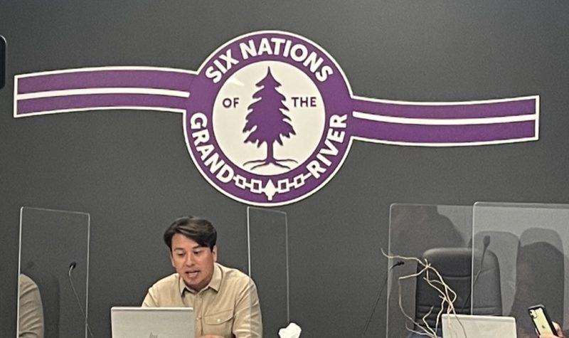 Council chambers at Six Nations. Man sits at desk with logo behind him on wall. dark grey wall purple and white logo on wall behind him.