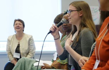 Une jeune dame parle dans un micro au milieu de dames assises.