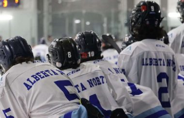 Hockey players on the bench, looking on the ice as they wait for their next shift. The game is indoors.