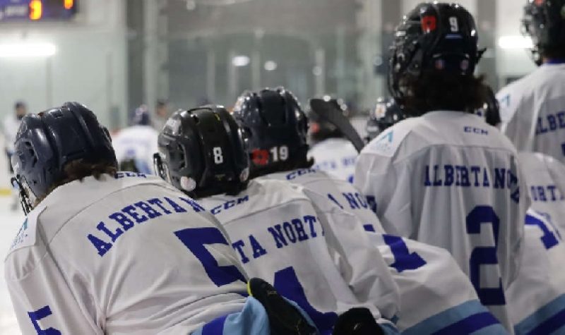 Hockey players on the bench, looking on the ice as they wait for their next shift. The game is indoors.