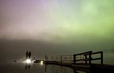 Two people stand at the edge of a dock on a giant lake with the northern lights dancing overhead. Time is nigh, weather is clear.