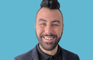 Pictured is a headshot of Quirion-Bouchard. He is standing against a baby blue background wearing a black suit.