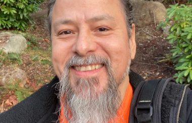 man with beard wearing orange shirt and black jacket smiles in outdoor setting