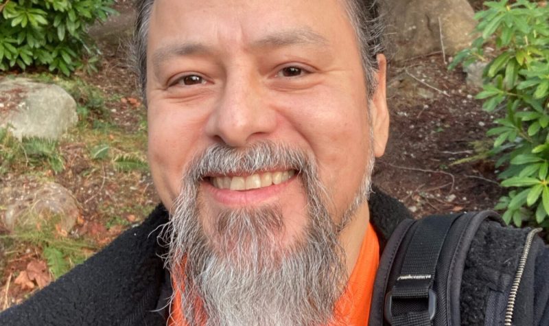 man with beard wearing orange shirt and black jacket smiles in outdoor setting