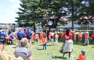 On aperçoit des dizaines d'enfants et quelque adultes sur le site du Festival du loup À Lafontaine. Plusieurs enfants portent les gilets oranges thématiques du festival. En arrière plan, on voit des arbres et un édifice municipal.