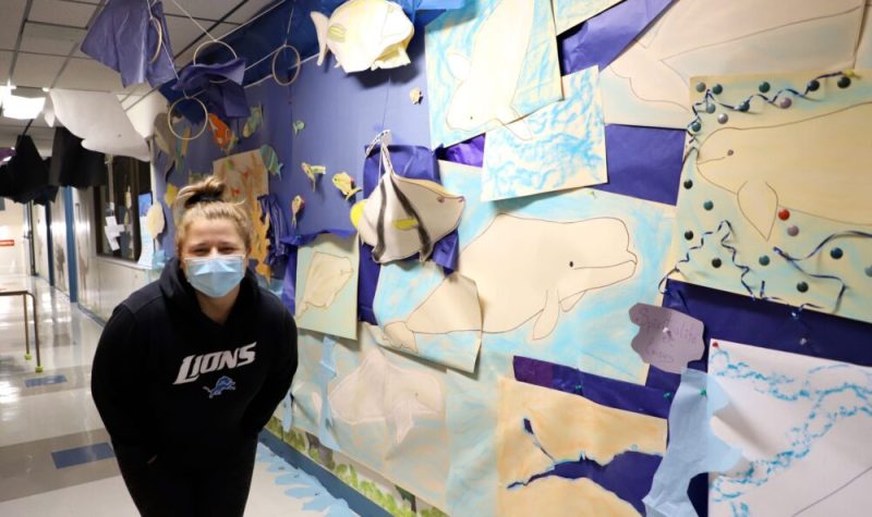 Une femme portant un masque de procédure pose aux côtés d’oeuvres en papier décorant le corridor d’une école.