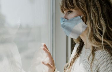 A woman with a medical face mask looks out the window. Photo by: Pxhere
