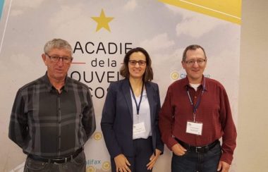 Deux hommes et une femme debout en avant d'une affiche avec l'écriteau Acadie de la Nouvelle-Écosse.