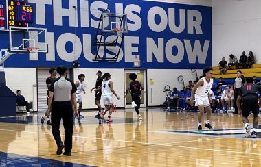 People in different basketball jerseys play on an indoor court with blue and white background.