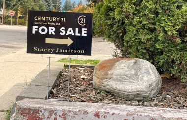 A For Sale sign sits in a roadside planter containing a rock and cedar bush.