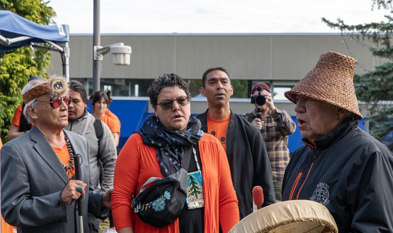 Image of four people, two wearing traditional regalia, standing outside wearing orange shirts while one of them drums on a hand drum.