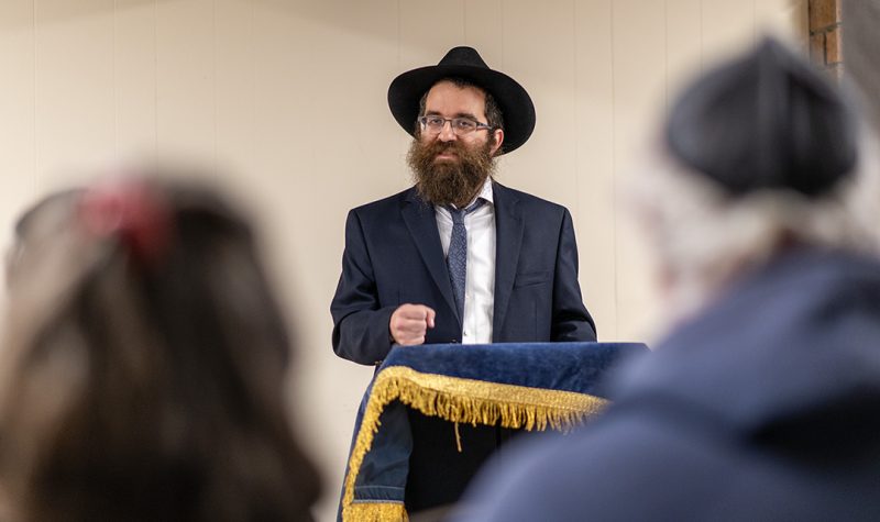 Rabbi Bentzi Shemtov speaks at a podium framed by two Jewish people in a shallow depth of field.