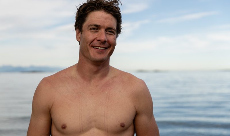 A smiling man with brown hair stands in front of the blue ocean with no shirt.
