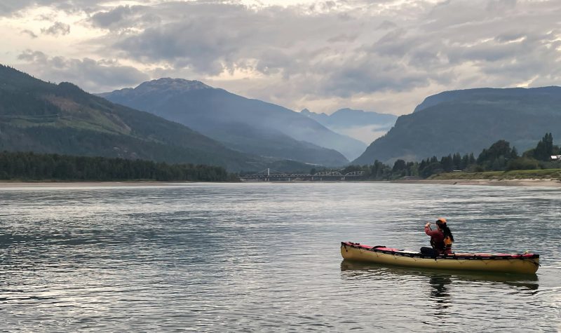 A person sits in a river in yellow canoe facing mountains and grey clouds.