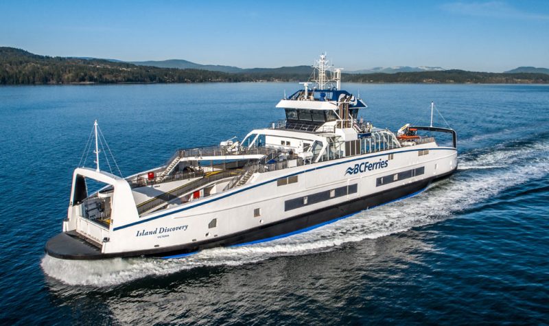 A large white and blue ferry boat plies through a calm sea with islands in the background.