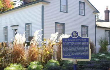 A white building with a blue and gold sign in front. There are large bushes of brown and green stems around the building.