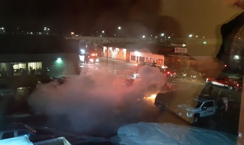 A dark night is lit up by the smoke and orange blaze of a vehicle fire outside of a hotel. Cars are on fire.