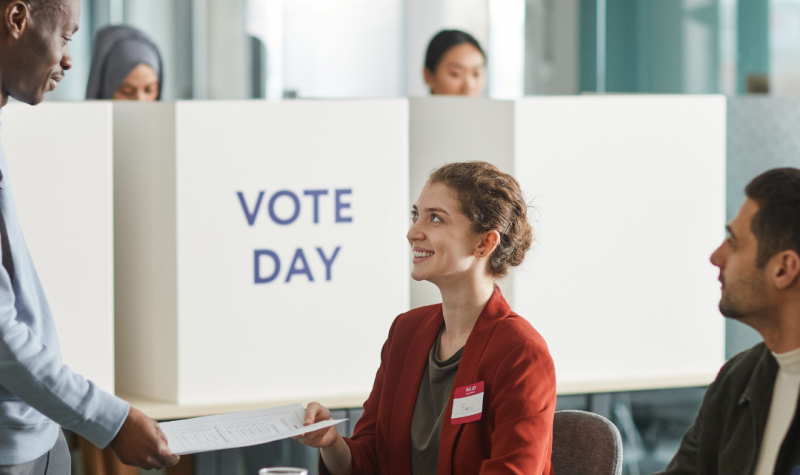 Un hombre se acerca a una mesa de votación donde están dos personas recibiendo su voto. Imagen ilustrativa vía Canva.com