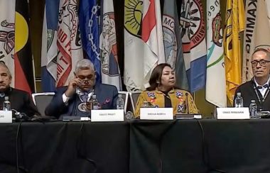 Six people sit at table covered with black cloth and name cards, First Nations flags in the background.