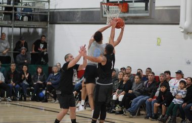 Three people jump at a basketball hoop with a crowd watching behind them just off the court.