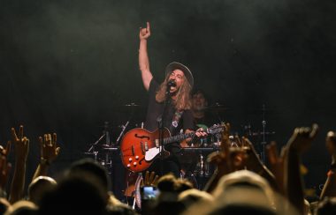 A crowd of people in the foreground raise their arms towards a man with a guitar singing on stage.