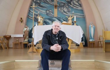 Un homme âgé assis dans l'escalier du choeur liturgique d'une petite église de village. On voit du bois et des couleurs pastels, du bleu et rose.