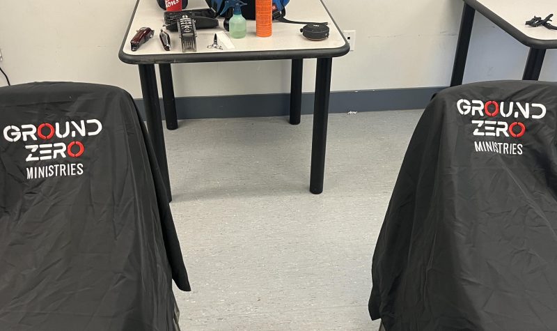 A room with two tables one table has bottles a blue backpack and some hair-cutting equipment and the other table is empty in this photo there are two chairs covered with two black cloths with the sign Ground Zero Ministries written on them
