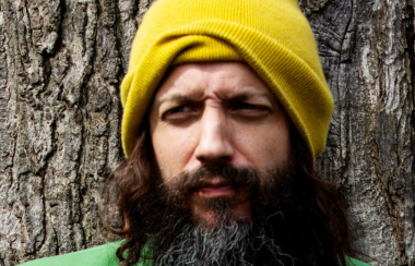 A person with a beard wearing a yellow beanie as they stand in front of a tree.