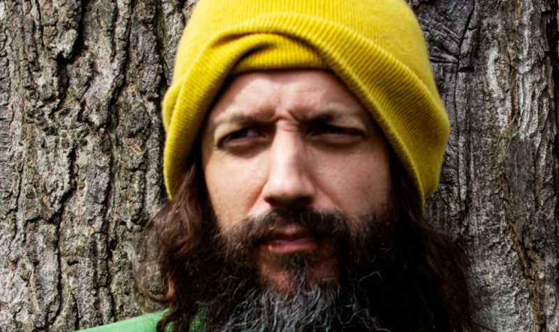 A person with a beard wearing a yellow beanie as they stand in front of a tree.