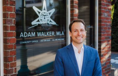 Photo of a man in a blue suit smiling in front of a window that reads Adam Walker, MLA