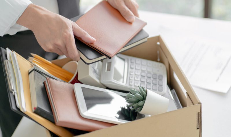 Une personne range des cahiers, une tablette électronique, une calculatrice et une plante dans une boîte.