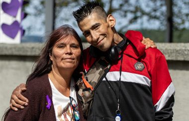 Photo of a Cree woman wearing a brown sweater with a purple ribbon and an Cree man wearing a red jacket smiling with their arms around each other.