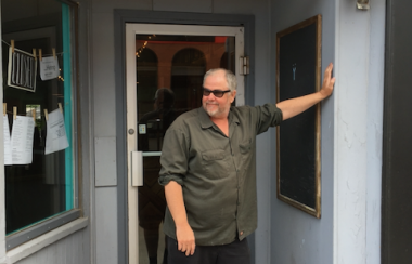 A man in sunglasses leaning against a doorway, with a sign above him that reads, Oysterhead.