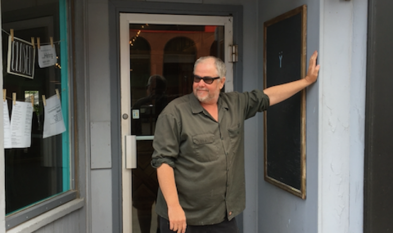 A man in sunglasses leaning against a doorway, with a sign above him that reads, Oysterhead.