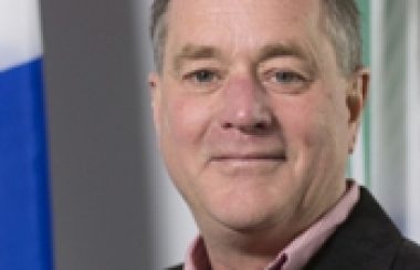 A professional headshot of Alain Gagnon wearing a dark suit and red shirt, standing in front of a Quebec flag.