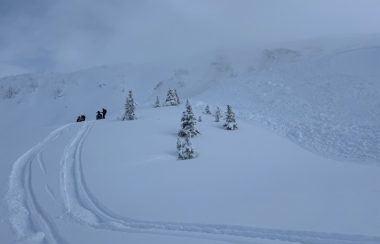 An avalanche on one side of a slop with snowmobilers on the ridge beside it.