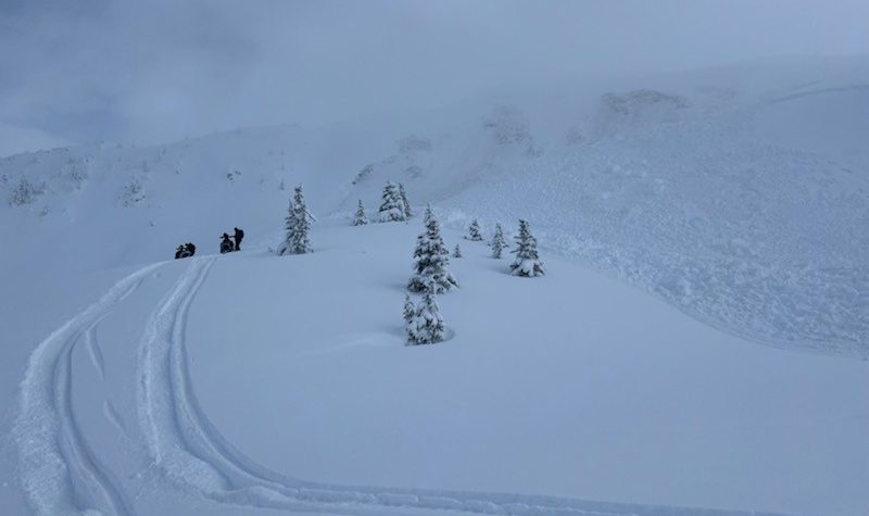 An avalanche on one side of a slop with snowmobilers on the ridge beside it.