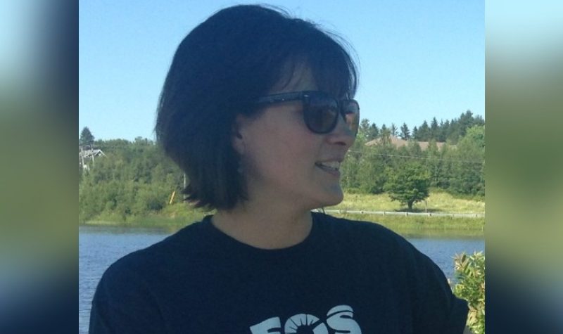 A woman with dark hair and sunglasses stands near a lake or other body of water under a blue sky wearing a black T-shirt with EOS written in white letters on the front. She is smiling and looking towards the right-hand side of the photo. There are trees and other greenery in the background beyond the water.