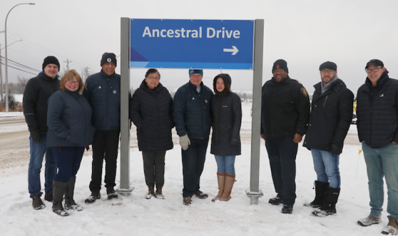 A group of people stand outside under and beside a street sign that reads Ancestral Drive.