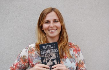 A young woman, Angelique Iles, poses in front of a pale wall holding a copy of her first published novel, Kentree's Stolen Souls--Escape into magic.