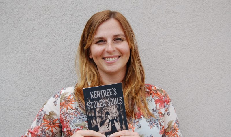 A young woman, Angelique Iles, poses in front of a pale wall holding a copy of her first published novel, Kentree's Stolen Souls--Escape into magic.