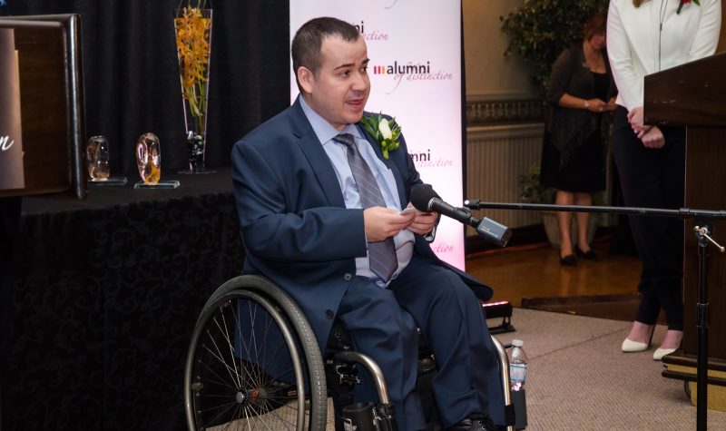 A person in a wheelchair is speaking into a microphone with people around them.