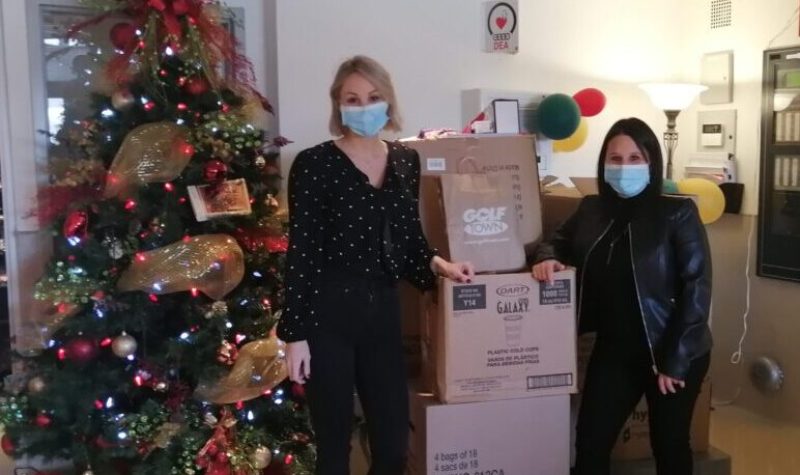 Deux femmes ( Audrey Gélinas à droite et Mélanie Rocco à gauche) avec un masque vêtues de noir se tiennent à côté d'un chariot rempli de boîtes de denrées. Un sapin de Noël se trouve dans la pièce.