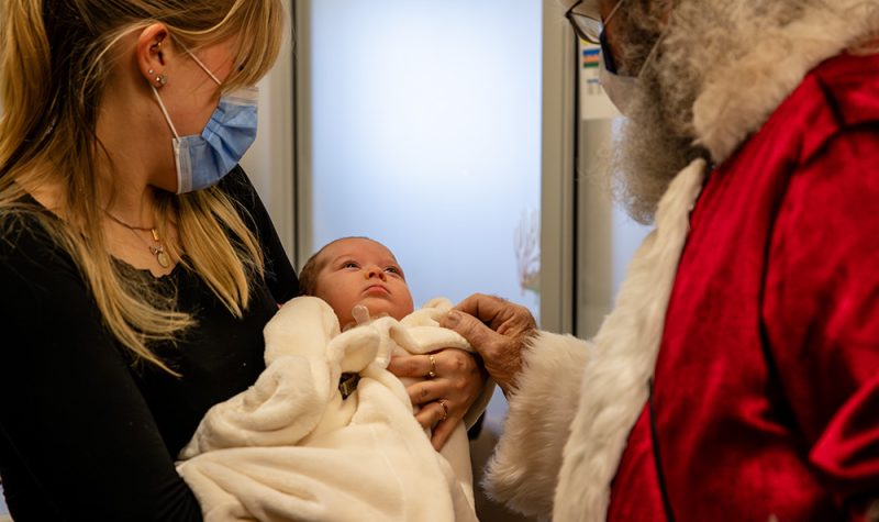 A man dressed as Santa Claus greets a baby held by her mother in a hospital with both adults wearing medical masks.