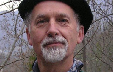 A man with a grey beard wearing a black hat stands in a rural forested area.