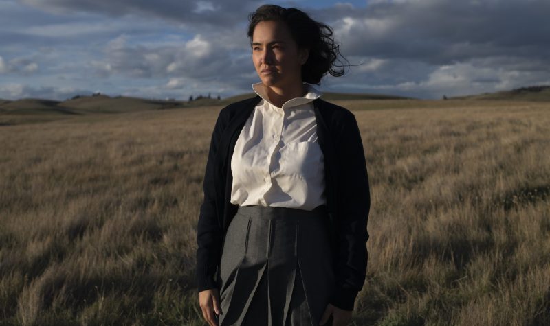 Aline Spears, played by Grace Dove, stands in the middle of a windy field looking off into the distance. Weather is partly cloudy.