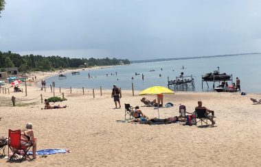 On aperçoit la plage de « Balm Beach » dans le canton de Tiny où des dizaines de vacanciers viennent profiter du soleil et de l'eau. On peut voir des vacanciers avec un parasol et des serviettes de plage tout le long de la plage de sable. En arrière plan on voit les boisés de Tiny.
