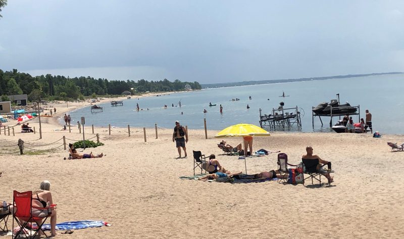 On aperçoit la plage de « Balm Beach » dans le canton de Tiny où des dizaines de vacanciers viennent profiter du soleil et de l'eau. On peut voir des vacanciers avec un parasol et des serviettes de plage tout le long de la plage de sable. En arrière plan on voit les boisés de Tiny.