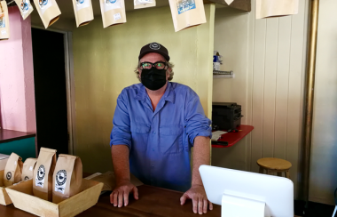 A man wearing a black face mask and blue t-shirt stands behind a cashier at a cafe.